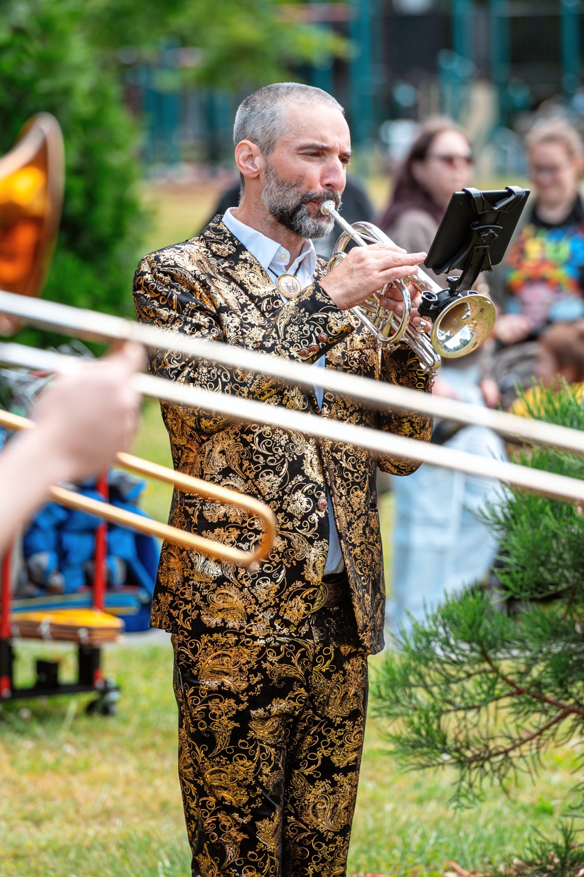 A musician in a dapper black and metallic gold patterned suit playing a coronet outside with nearby trombone slides out-of-focus in the foreground