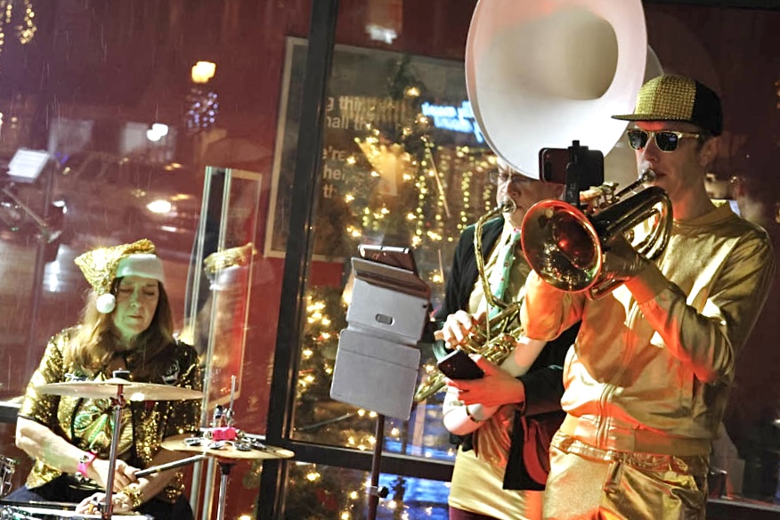 People wearing shiny metallic gold playing brass and percussion instruments at night around the holidays.